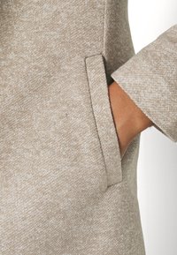 Béžová texturovaná látka s jemným vzorem, obsahující boční kapsu a otevřený manžet. Detail materiálu a ruky.