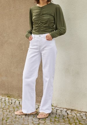 Osoba nosząca oliwkowozielony marszczony top z długim rękawem, białe szerokie spodnie z wysokim stanem oraz beżowe plecione sandały wsuwane stoi na brukowanej nawierzchni.