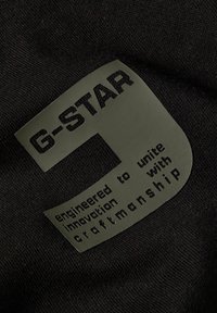 Tela negra con una etiqueta texturizada verde que presenta "G-STAR" y el texto "diseñado para unir innovación con artesanía."