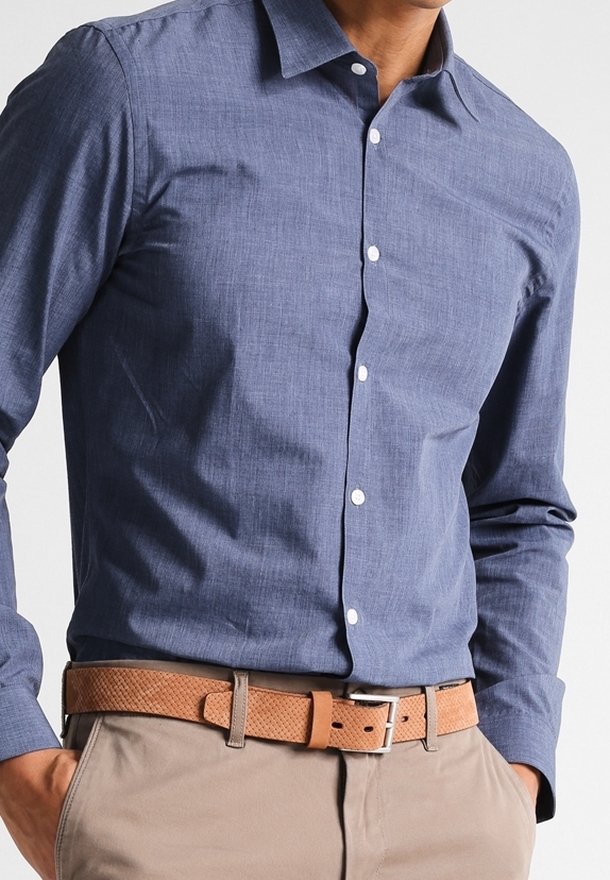 Homme portant une chemise bleue boutonnée rentrée dans un pantalon beige avec une ceinture marron texturée, les mains dans les poches.