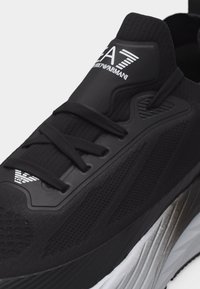 Μαύρο αθλητικό παπούτσι EA7 Emporio Armani με πλεκτό επάνω μέρος, μαύρα κορδόνια και λευκό λογότυπο στη γλώσσα και κοντά στη μύτη, καθώς και ασημί ανάγλυφη σόλα.