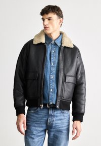 Black leather bomber jacket with a beige shearling collar, front pockets, ribbed cuffs, and zip closure. Worn over a blue denim shirt.