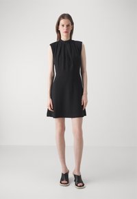 Black sleeveless dress with a high neckline and pleated detail, featuring a fitted bodice and flared skirt. Paired with black slide sandals.