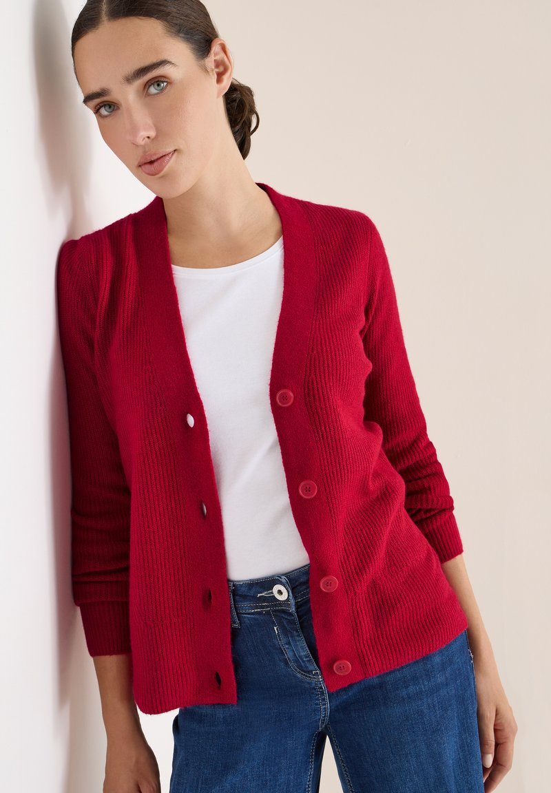 Rote gerippte Strickjacke mit V-Ausschnitt und Knöpfen, getragen über einem weißen Hemd. Hat lange Ärmel und wird mit blauen Jeans kombiniert.