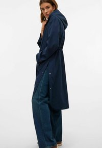 Marinblå vattentät jacka med huva, åtdragen midja, tryckknappar och sidofickor. Buren över vida denimbyxor. Slät yta.