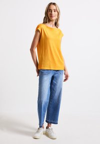 Hořčicově žlutý top s krátkým rukávem spárovaný se světle modrými širokými džíny. Outfit doplňují bílé tenisky na neutrálním pozadí.