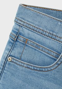 Los jeans de denim azul claro presentan una cintura alta, costuras visibles en un color mostaza que contrasta, y un pequeño bolsillo delantero con un acento metálico.