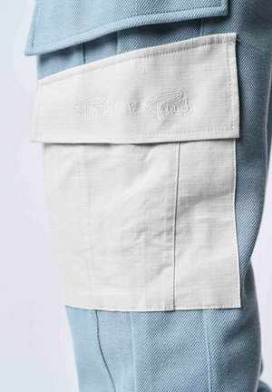 Pantalón de tejido azul texturizado con un bolsillo de parche blanco que presenta el texto bordado "Project x Paris", resaltando el diseño único de capas.