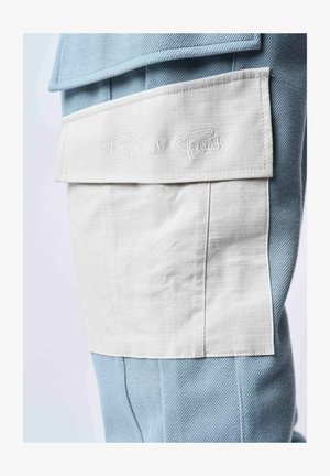 Pantaloni in tessuto blu con texture, dotati di una tasca a toppa bianca con la scritta ricamata "Project x Paris", evidenziando un design di sovrapposizione unico.