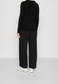 Svart, långärmad tröja i mjukt material, tillsammans med löst sittande svarta byxor. Vita sneakers med grå detaljer syns.