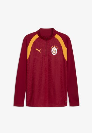 Haut de sport à manches longues bordeaux avec des accents jaunes aux épaules, une fermeture éclair sur le quart, logo Puma et blason d’un club de football avec quatre étoiles.