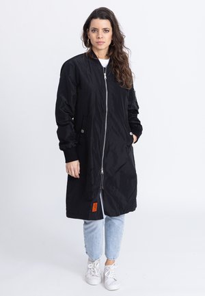 Schwarze lange Jacke aus glänzendem Material, mit einem Frontreißverschluss, Seitentaschen und einem orangefarbenen Etikett am Saum. Über hellen Jeans getragen.