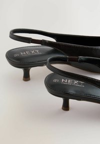 Svarta slingback-klackar med blanka ytor och låg klack, med märket "NEXT" på innersulan. Enkelt, elegant design.