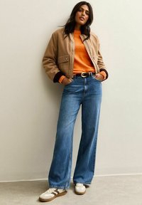Femme portant une veste beige, un pull orange, un jean bleu large, des baskets blanches, se tenant les mains dans les poches contre un mur uni.