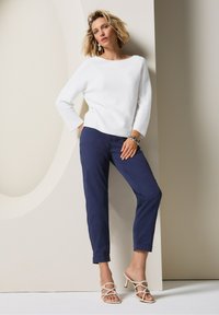 Witte, lange mouw trui met een bootnek, gecombineerd met marineblauwe, slim-fit broek. Beige haksandalen. Eenvoudige achtergrond met gedempte tinten.