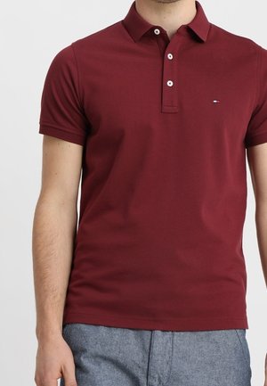 Homme portant un polo bordeaux à manches courtes avec des boutons blancs et un pantalon gris, debout devant un fond uni.