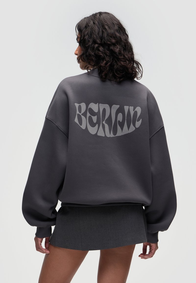 Femme aux cheveux bouclés et foncés vue de dos portant un sweat-shirt gris foncé surdimensionné avec "BERLIN" dans le dos et une jupe courte foncée.