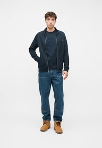 Νέος άνδρας με σγουρά μαλλιά, φορώντας μια μπλε μπλούζα με φερμουάρ, σκούρο μπλε μπλουζάκι, άνετο τζιν με τσέπες με επιδιορθώσεις και καφέ εργασιακές μπότες, όρθιος.