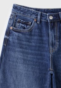 Jean denim bleu foncé avec poches avant, passants de ceinture, surpiqûres contrastantes et fermeture par bouton métallique sur un fond clair.