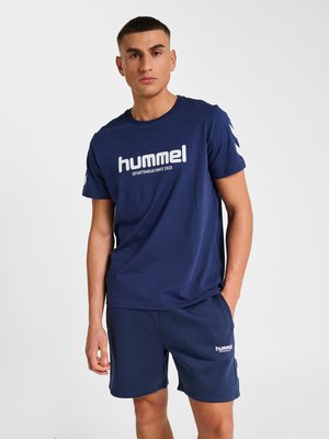 Mand iført marineblå hummel t-shirt og shorts, stående med den ene hånd i lommen mod en ensfarvet hvid baggrund.