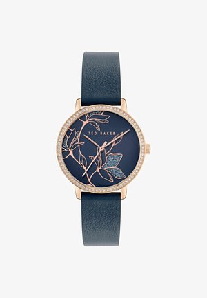 Hodinky v ružovo-zlatom tóne s tmavomodrým koženým remienkom, kvetinovým vzorom na ciferníku a korunkou pokrytou kryštálmi. Značka Ted Baker.
