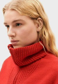 Pull en tricot rouge avec un col montant à côtes et fermeture éclair. Présente un motif texturé le long de l'encolure. Apparence de tissu doux.