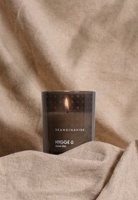 Bocal de bougie en verre ambre avec une surface texturée, arborant le nom de la marque "SKANDINAVISK" et l'étiquette "HYGGE" ; bougie allumée à l'intérieur.