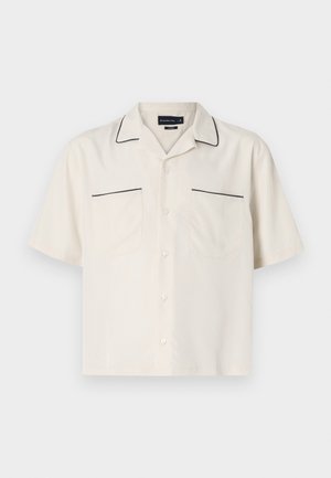 Bež srajca s kratkimi rokavi z črnimi obrobami ob ovratniku in robovih žepov. Ima dva sprednja žepa in gumbasto zapiranje.