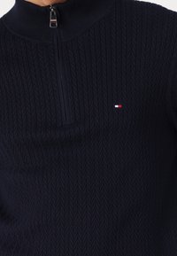 Marineblauer Strickpullover mit halb durchgehendem Reißverschluss, im strukturierten Fischgrätenmuster und einem kleinen Logo in Rot, Weiß und Blau auf der Brust.