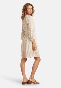 Beiges Kleid mit Zickzackmuster, langen Ärmeln und einer taillierten Taille. Kombiniert mit braunen Riemchensandalen steht das Modell in einer entspannten Pose.
