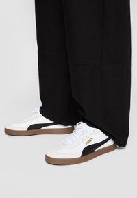 Vita sneakers med svarta accenter, med en texturerad sula och en enkel snörningsdesign. Varumärke i guld på sidan. Svarta byxor täcker en del av skorna.