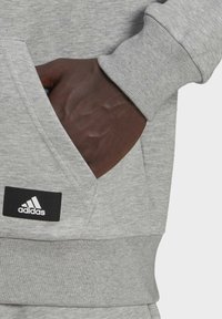 Felpa grigia con tasca frontale, polsini e orlo a costine. Presenta un logo Adidas su un'etichetta nera sopra la tasca. Tessuto dalla trama liscia.