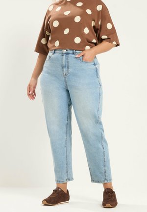 Personne portant un jean taille haute bleu clair, une chemise marron à pois et des baskets marron, debout avec une main dans la poche.