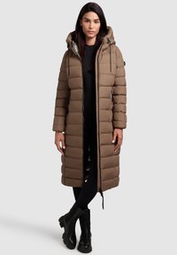 Lange bruine gewatteerde jas met capuchon, voorzien van een doorgestikt patroon, ritssluiting aan de voorkant, zijzakken en zwarte accenten. Draagt over zwarte leggings en laarzen.