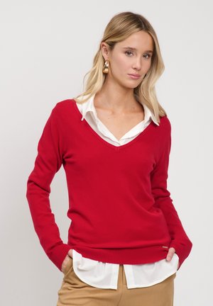 Lyshåret kvinde iført guldbøjleøreringe, en rød V-hals sweater over en hvid skjorte og beige bukser med hænderne i lommerne mod en hvid baggrund.