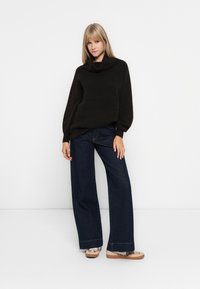 Übergroßer dunkelbrauner Strickpullover mit einem Loopkragen, kombiniert mit weit geschnittenen, dunkelblauen Jeans. Die Schuhe haben eine beigen Basis mit einem gemusterten Design.
