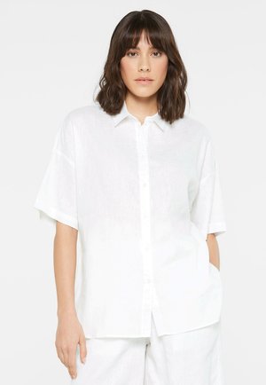 Woman with medium-length brown hair stands facing forward, wearing a loose-fitting white short-sleeve button-up shirt and matching pants.