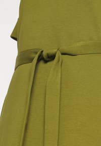Gros plan sur une robe en tissu vert olive avec une ceinture nouée autour de la taille, montrant la texture et les détails de couture.