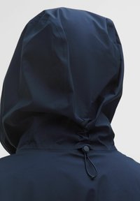 Üksikasjalik vaade tumesinise vihmakeebi kapuutsi, mille taga on reguleeritav nöör ja tõkistik, heledal taustal.