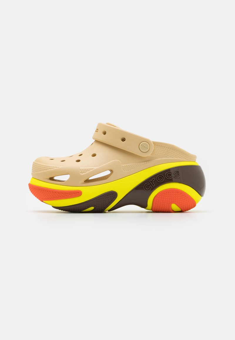 Sabot din plastic bej deschis, cu decupaje, accente galbene și portocalii, o talpă maro și un bretele pe spate, având logo-ul "CROCS".