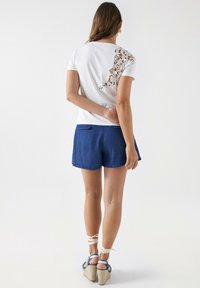 Wit T-shirt met kantdetails op één schouder, gecombineerd met blauwe denim shorts. Sleehakken met enkellinten. Soepele stoftextuur.