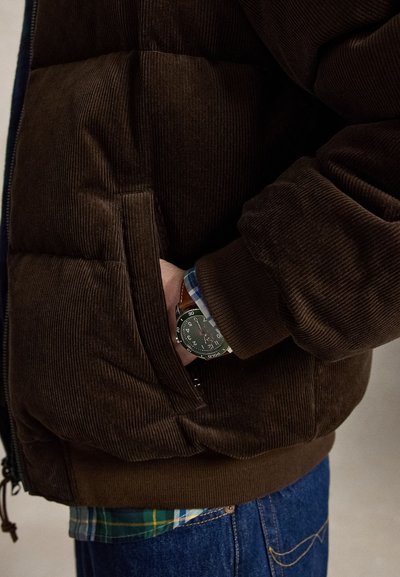 Άτομο που φοράει καφέ μπουφάν από κορδούρα με το χέρι στην τσέπη, δείχνοντας ένα ρολόι με καφέ δερμάτινο λουράκι και μπλε-πράσινο καντράν.