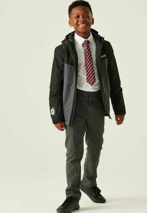 Uśmiechnięty chłopiec w mundurku szkolnym, ubrany w czarno-szarą kurtkę z kapturem, białą koszulę, paskowany krawat, szare spodnie i czarne buty, stojący.