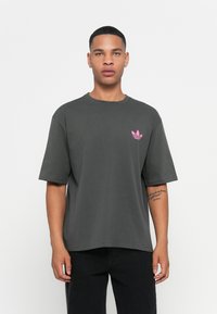 Giovane uomo con capelli corti, che indossa una maglietta larga grigio scuro con un piccolo logo rosa sul lato sinistro del petto e pantaloni neri, in piedi su uno sfondo bianco.