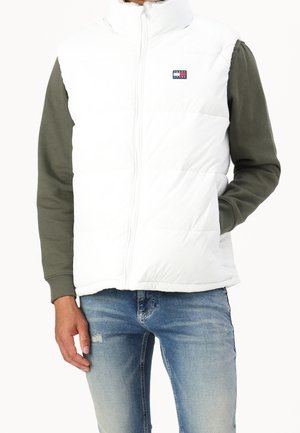 Personne portant un gilet matelassé blanc sans manches sur une chemise olive à manches longues et un jean bleu clair déchiré, debout avec une main dans la poche.