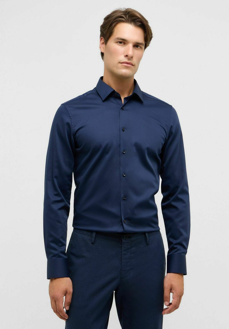 Man staat in een aansluitend donkerblauw overhemd met lange mouwen en bijpassende broek tegen een witte achtergrond.