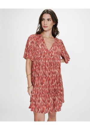 Femme aux longs cheveux bruns portant une robe ample courte rouge et blanche à motifs avec des manches papillon, debout devant un fond blanc uni.