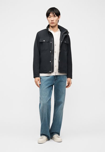 Νέος άνδρας με μαύρο σακάκι με κουμπιά, μπεζ ζακέτα, μπλε τζιν και λευκά αθλητικά παπούτσια, στέκεται μπροστά σε απλό λευκό φόντο.