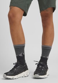 Scarpe da ginnastica nere con suole bianche, calzini grigi a fantasia con disegni di montagne, abbinati a pantaloni corti verdi. Texture lisce visibili.