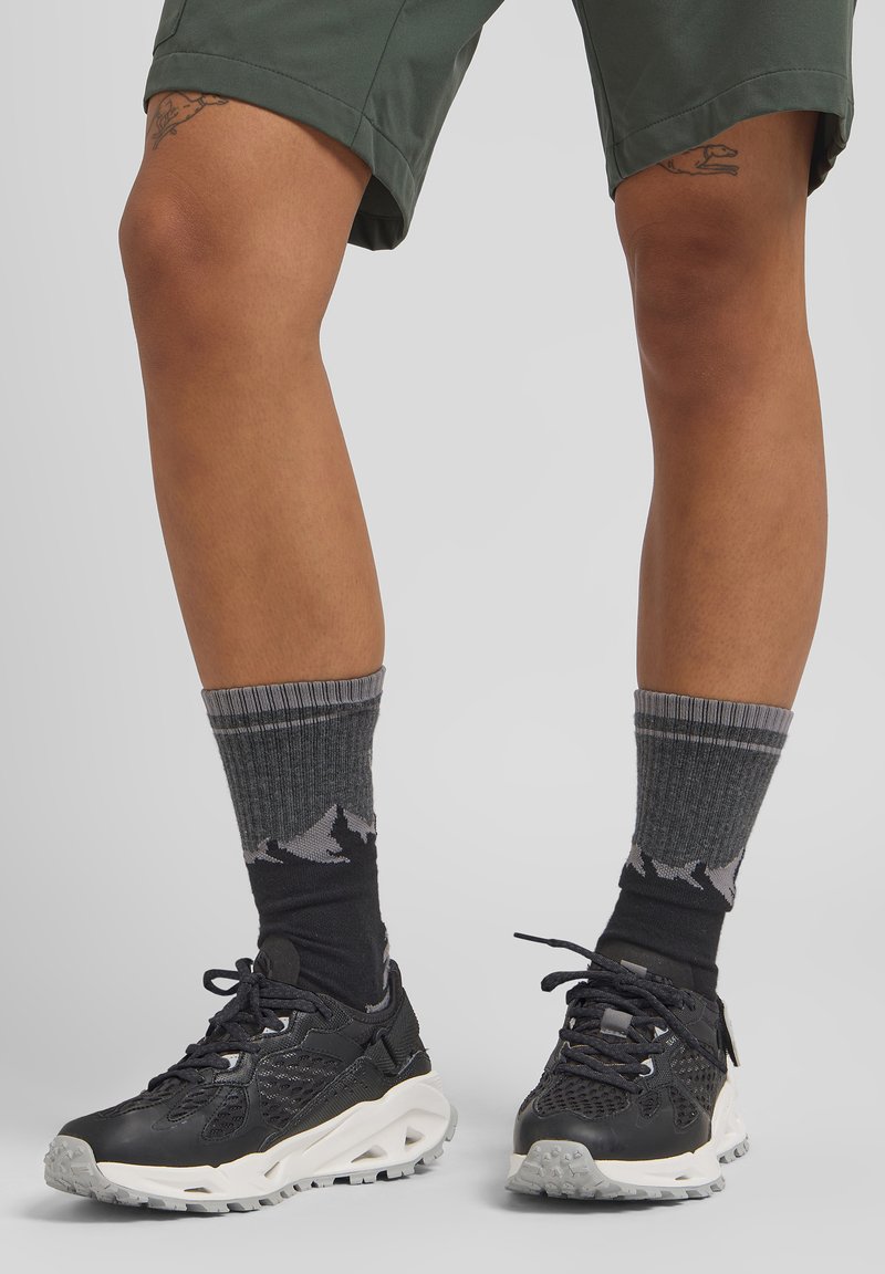 Scarpe da ginnastica nere con suole bianche, calzini grigi a fantasia con disegni di montagne, abbinati a pantaloni corti verdi. Texture lisce visibili.